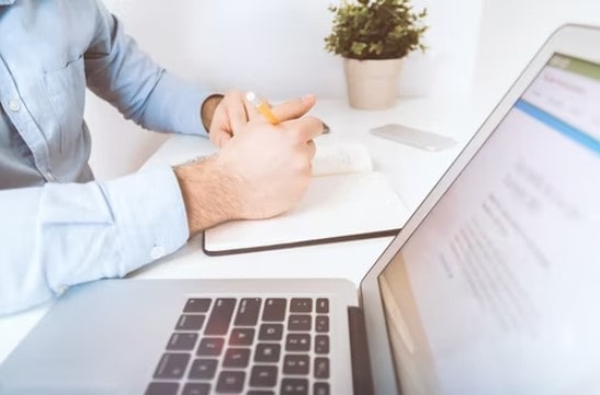 A man making bookkeeping notes in a notebook next to a laptop not knowing the four advantages of outsourcing his bookkeeping.